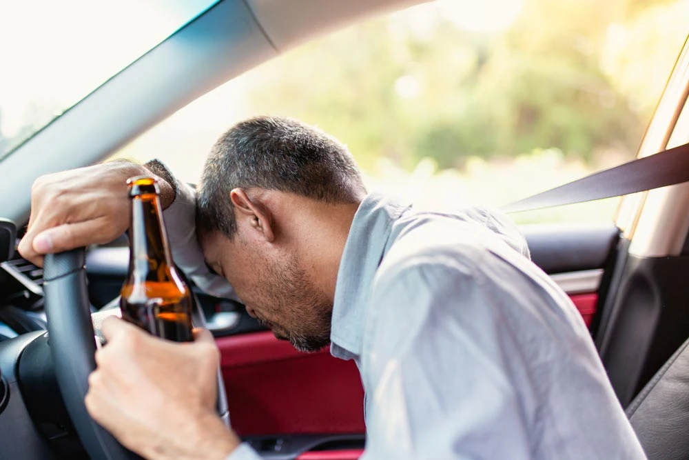 Drunk asian young man drives a car with a bottle of beer with sunset background, Dangerous driving concept